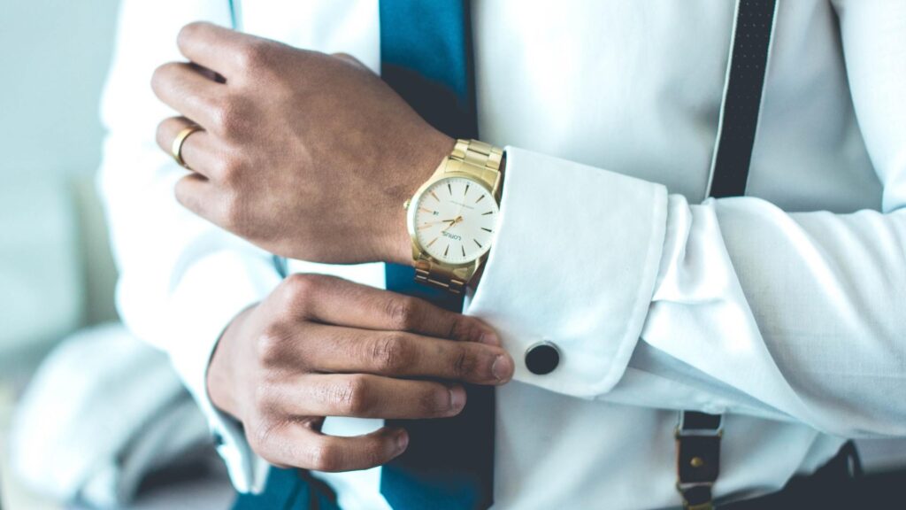 Una persona abrochándose el puño de la camisa y mostrando un reloj analógico para hombre dorado. El hombre lleva corbata azul y tirantes sobre una camisa blanca.