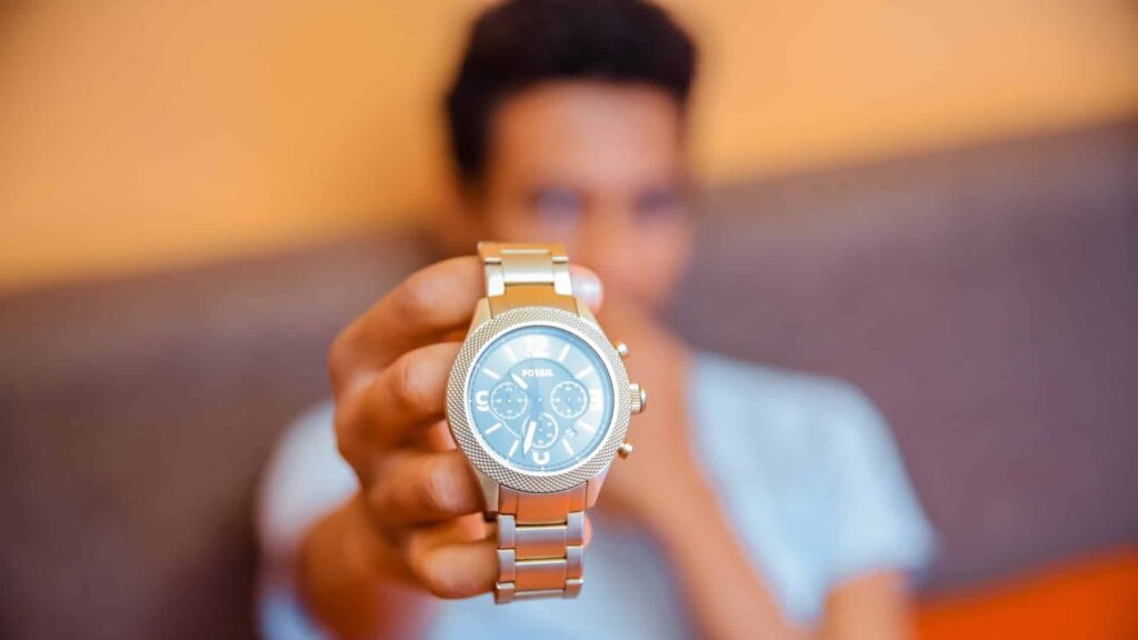 Un chico mostrando un reloj de pulsera en su mano. El reloj tiene una correa de metal y una esfera azul con números blancos. El fondo está desenfocado.