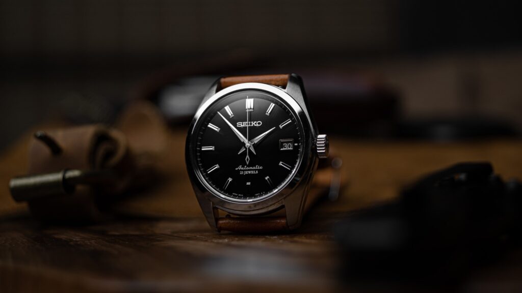 Reloj Seiko de esfera negra y correa de cuero marrón sobre una superficie de madera. En el reloj se puede leer “Seiko Automatic 23 Jewels”.