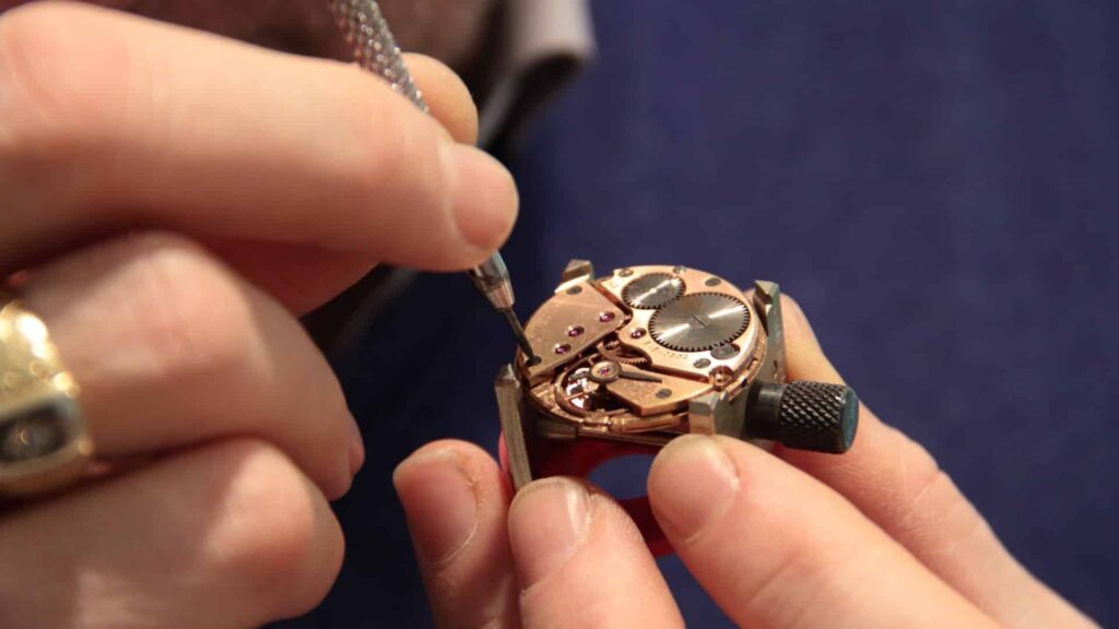 Un relojero trabajando en un reloj de pulsera de oro. El fondo es un material de terciopelo azul. Las manos del relojero son visibles en primer plano.